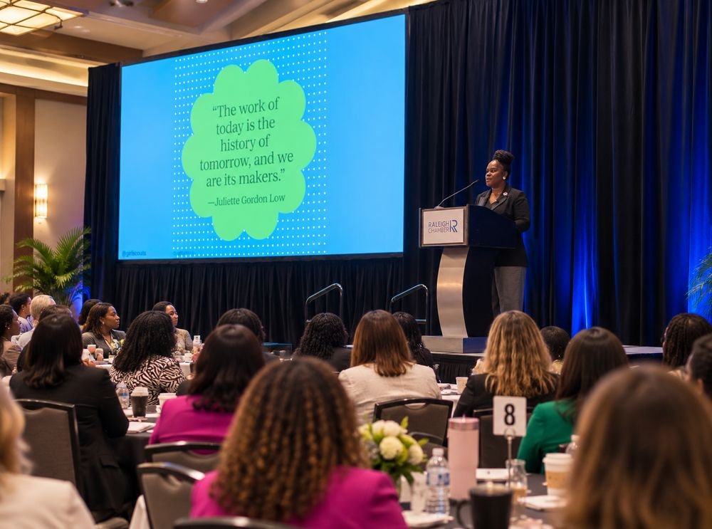 Latisha Newby speaking from the podium at a Raleigh Chamber event, to a full room seated at round tables.